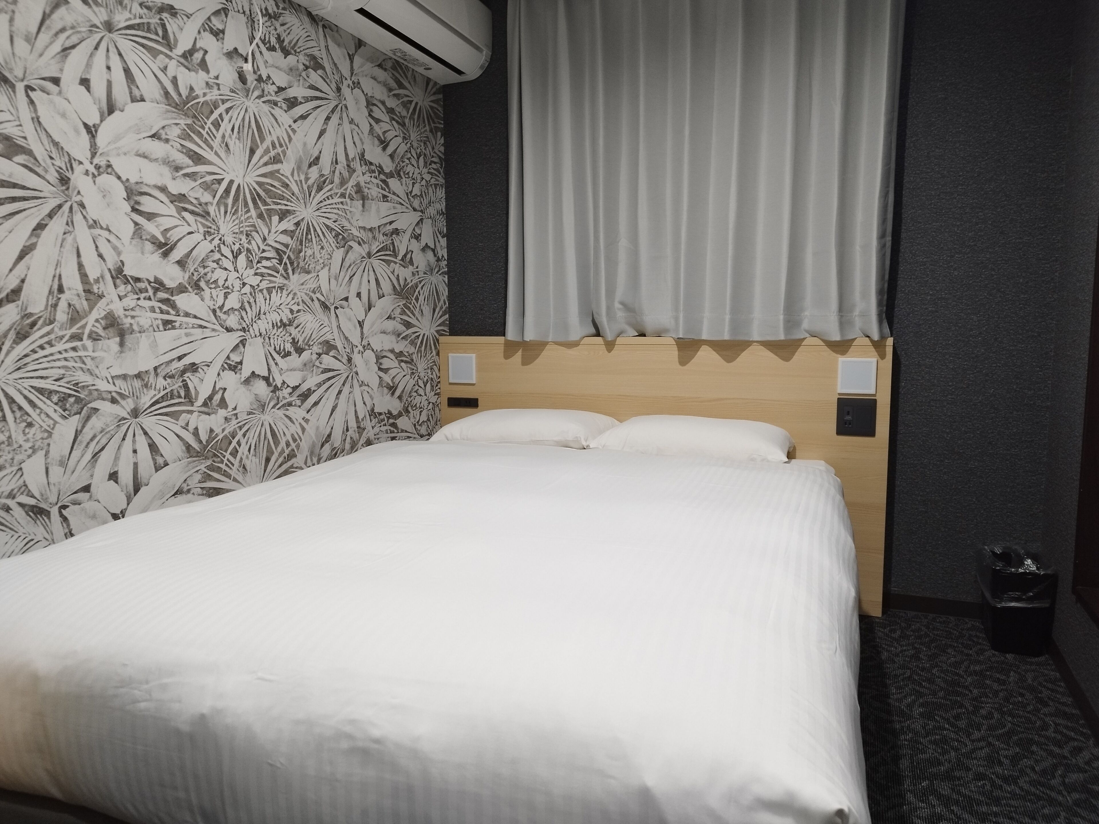 Une chambre d’hôtel avec un lit, un papier peint à motifs floraux, un rideau gris et un climatiseur fixé au mur.