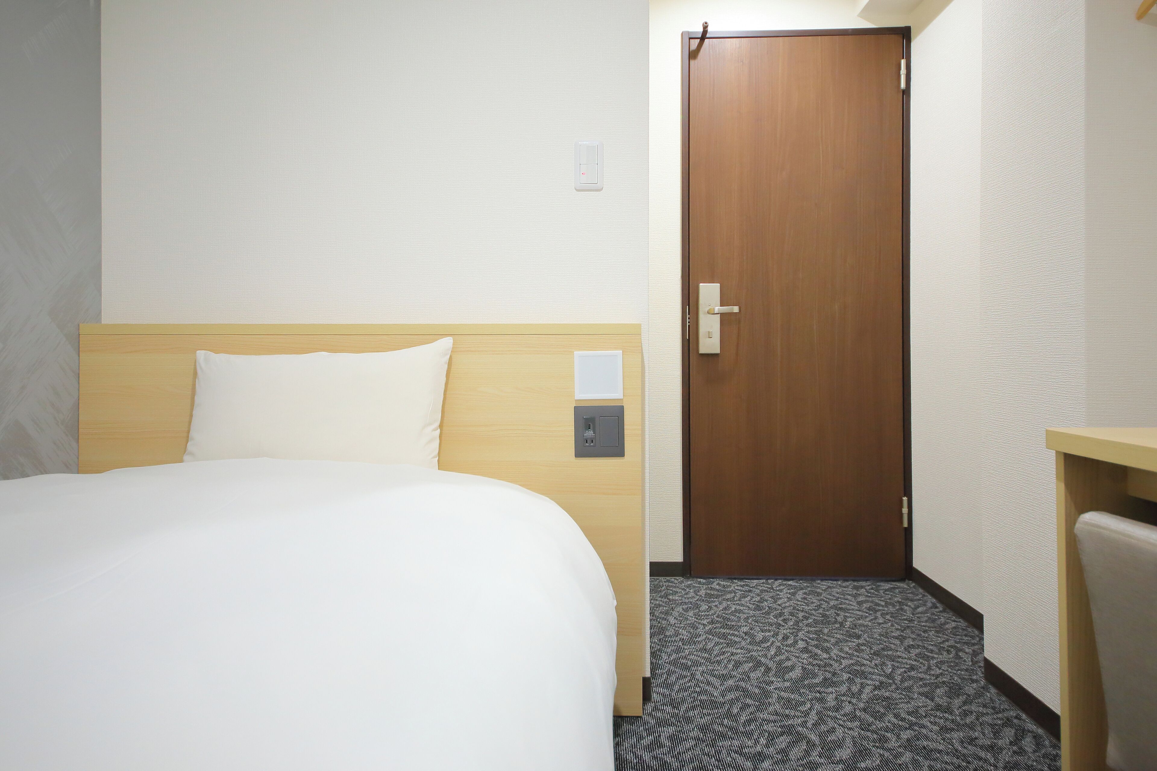 A hotel room with a bed, a wooden headboard, a door, and a small desk with a chair.