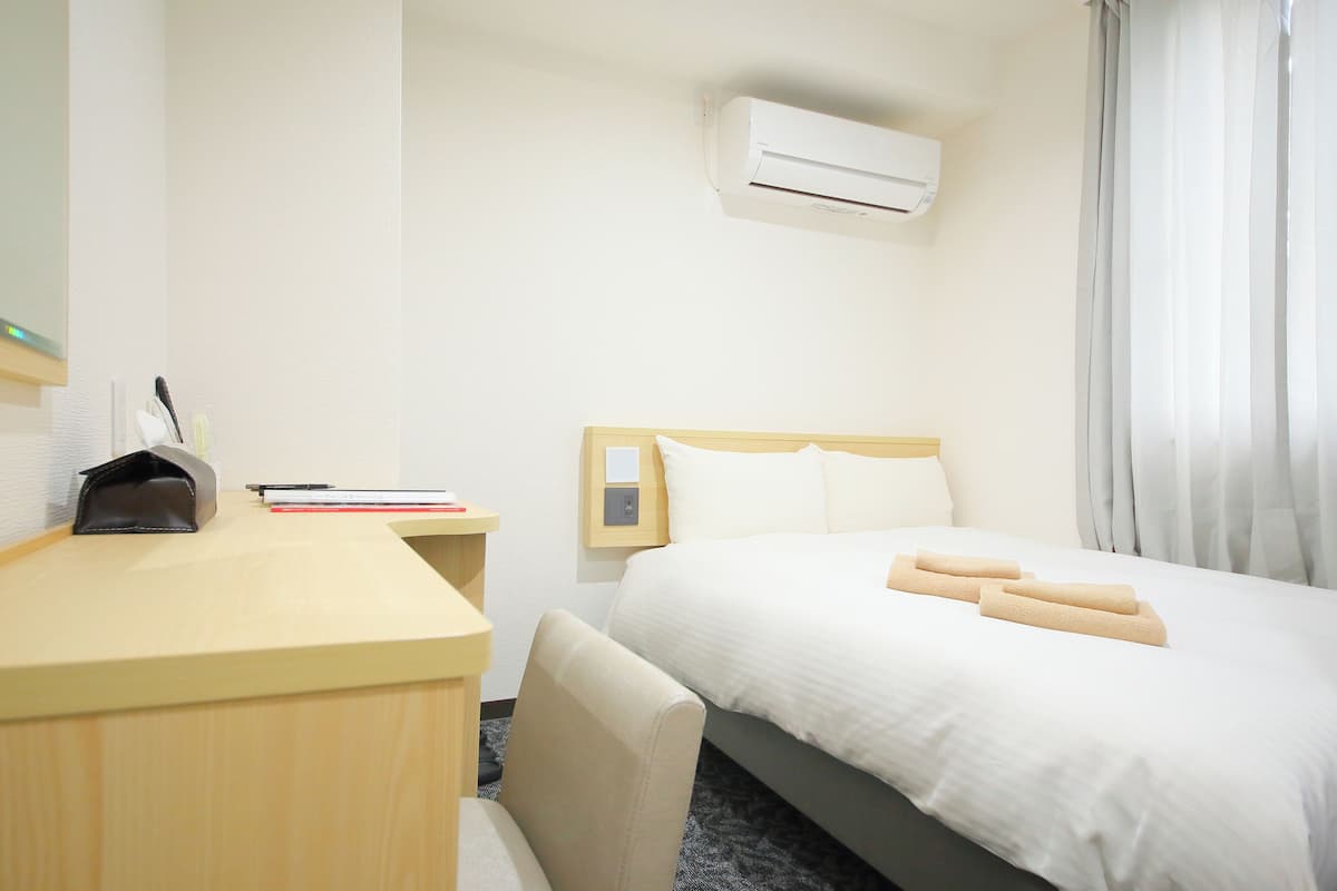 Habitación de hotel con cama, escritorio, silla, mesita de noche, aire acondicionado y ventana con cortinas.
