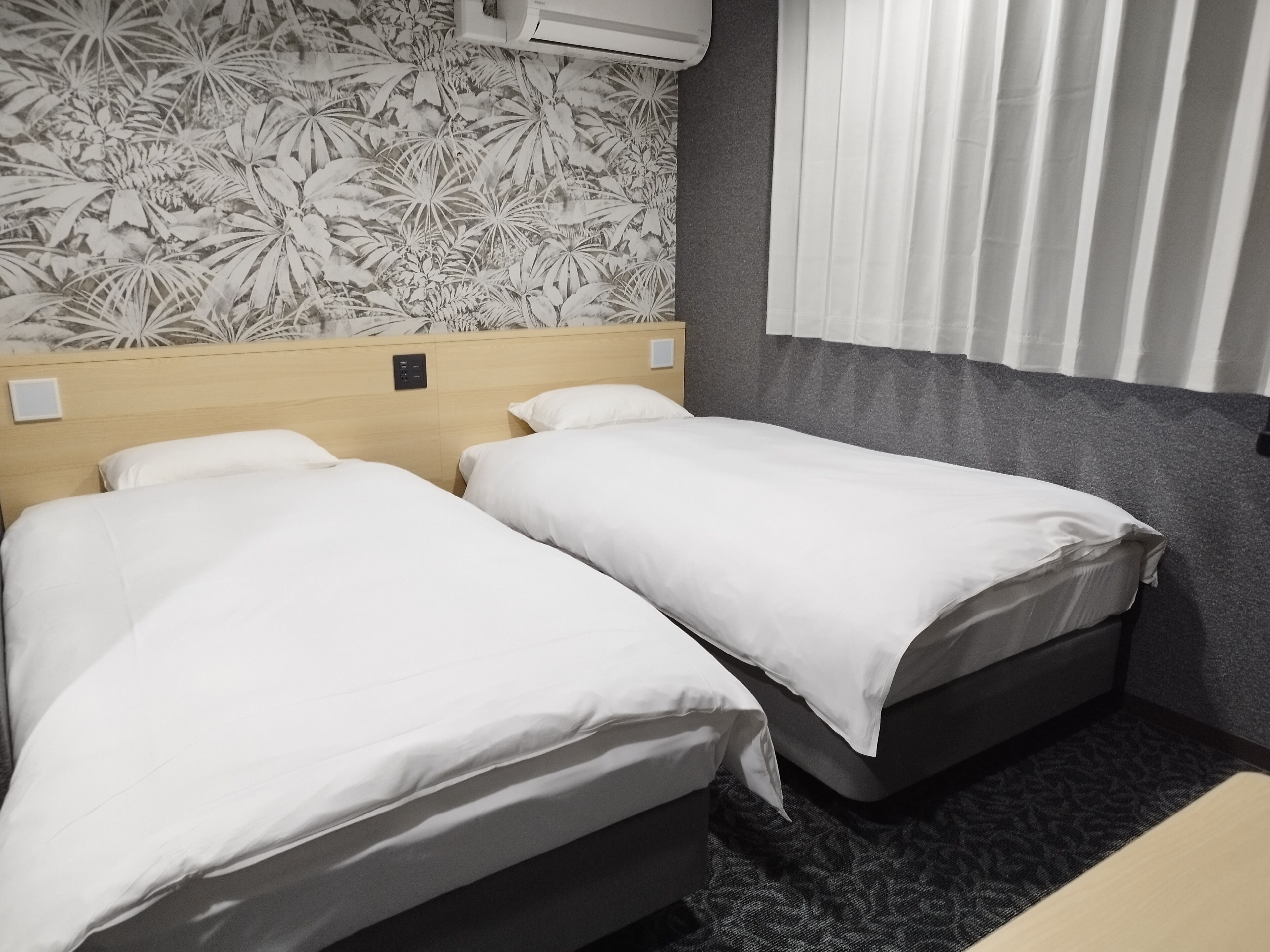 A hotel room with two beds, a headboard with a light switch, a wall air conditioner, and a window with blinds.