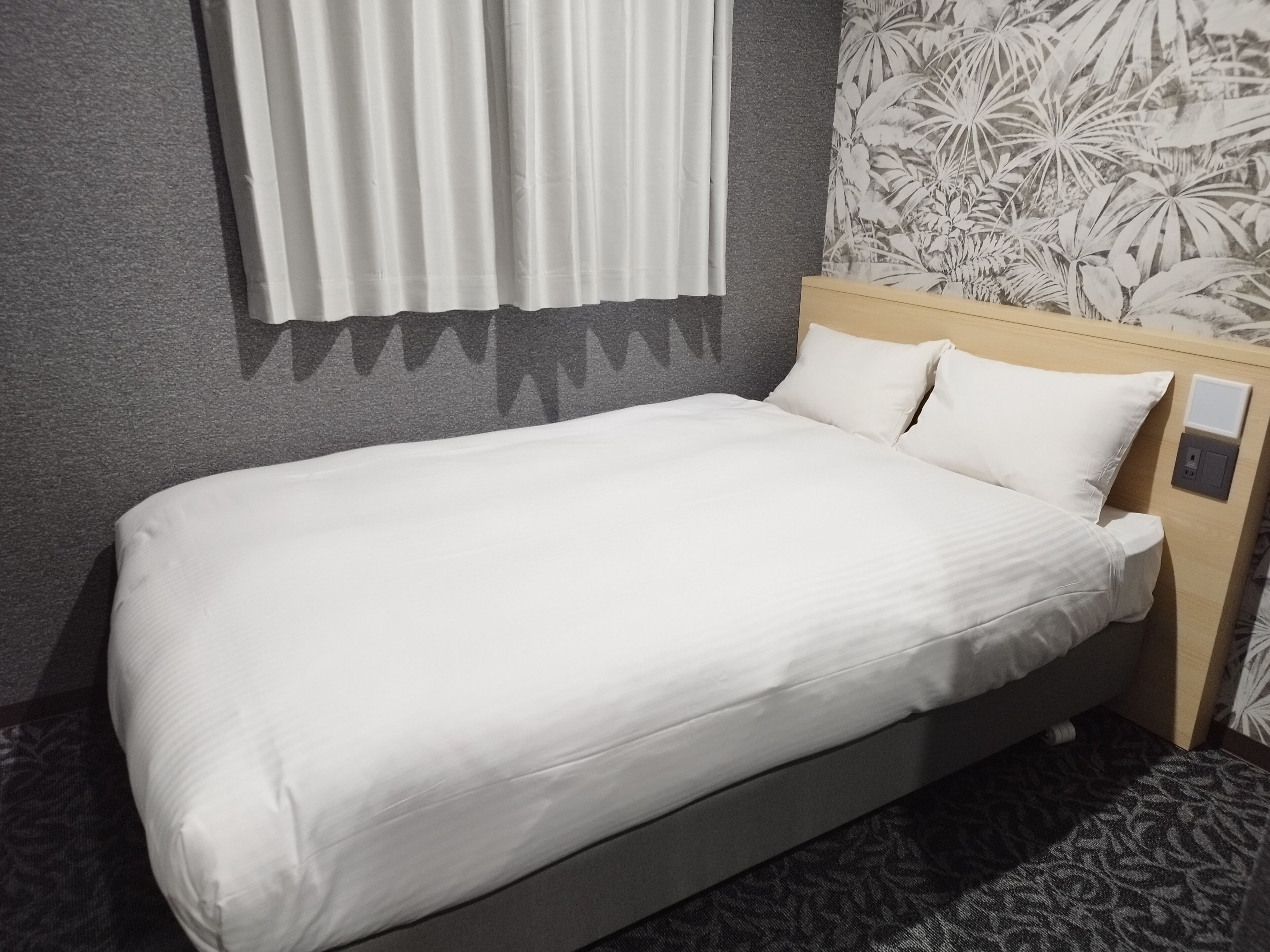 A neatly made bed with white linens in a room with a patterned wallpaper and a wooden headboard.