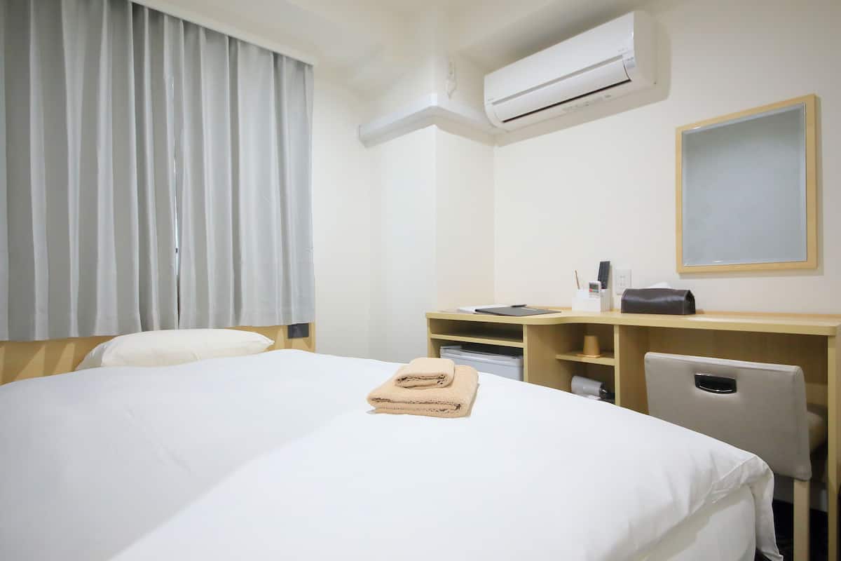 Una habitación de hotel con cama, escritorio, silla, aire acondicionado y ventana con cortinas.