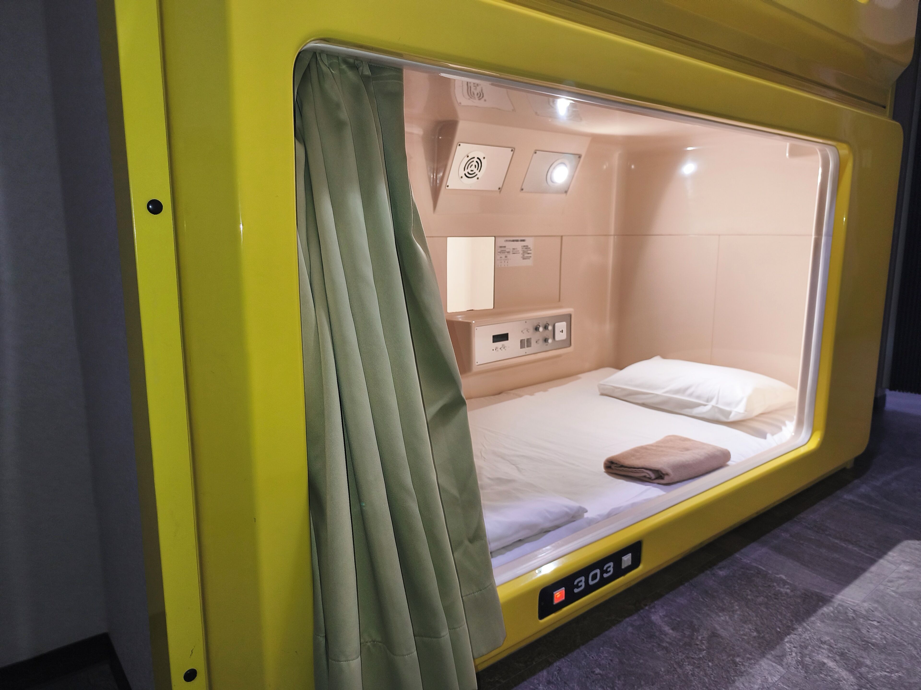 A yellow sleeper train compartment with a single bed, a control panel, and a window with curtains.