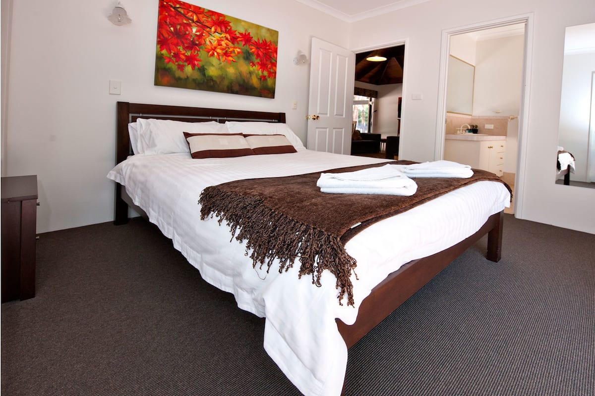 A neatly made bed with white linens and a brown throw, a painting of red flowers on the wall, and a bathroom visible through an open door.
