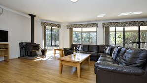 A living room with a wooden floor, a black leather sofa, a wooden coffee table, a fireplace, and large windows with curtains. - Margaret River Hideaway (Bramley)