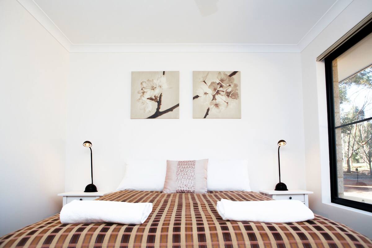 A bedroom with a large bed, two bedside tables, two wall-mounted lamps, a window with a view of trees, and two wall art pieces.