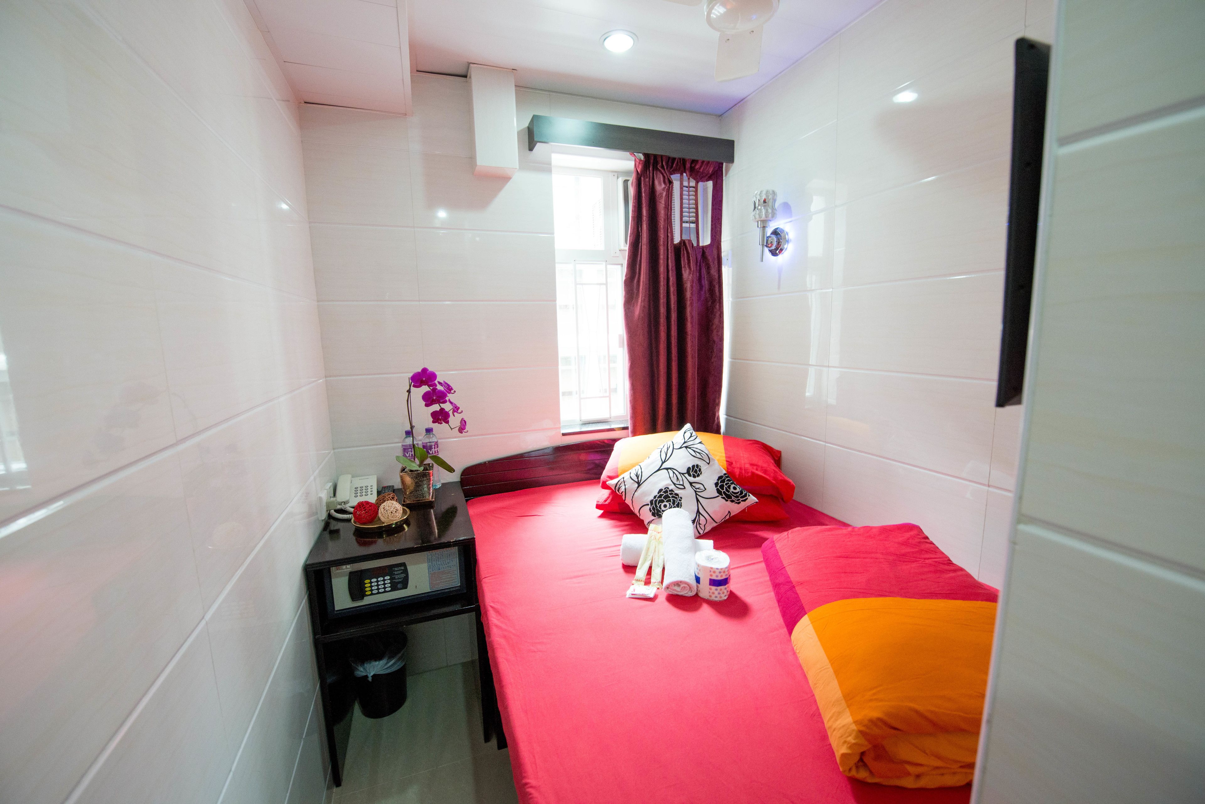 A small, brightly lit room with a bed, a bedside table with a phone, and a window with curtains.