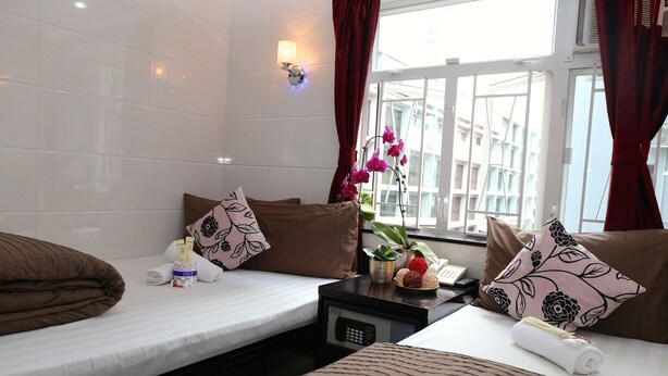 A hotel room with two beds, a bedside table with a lamp, a window with curtains, and a view of buildings outside.