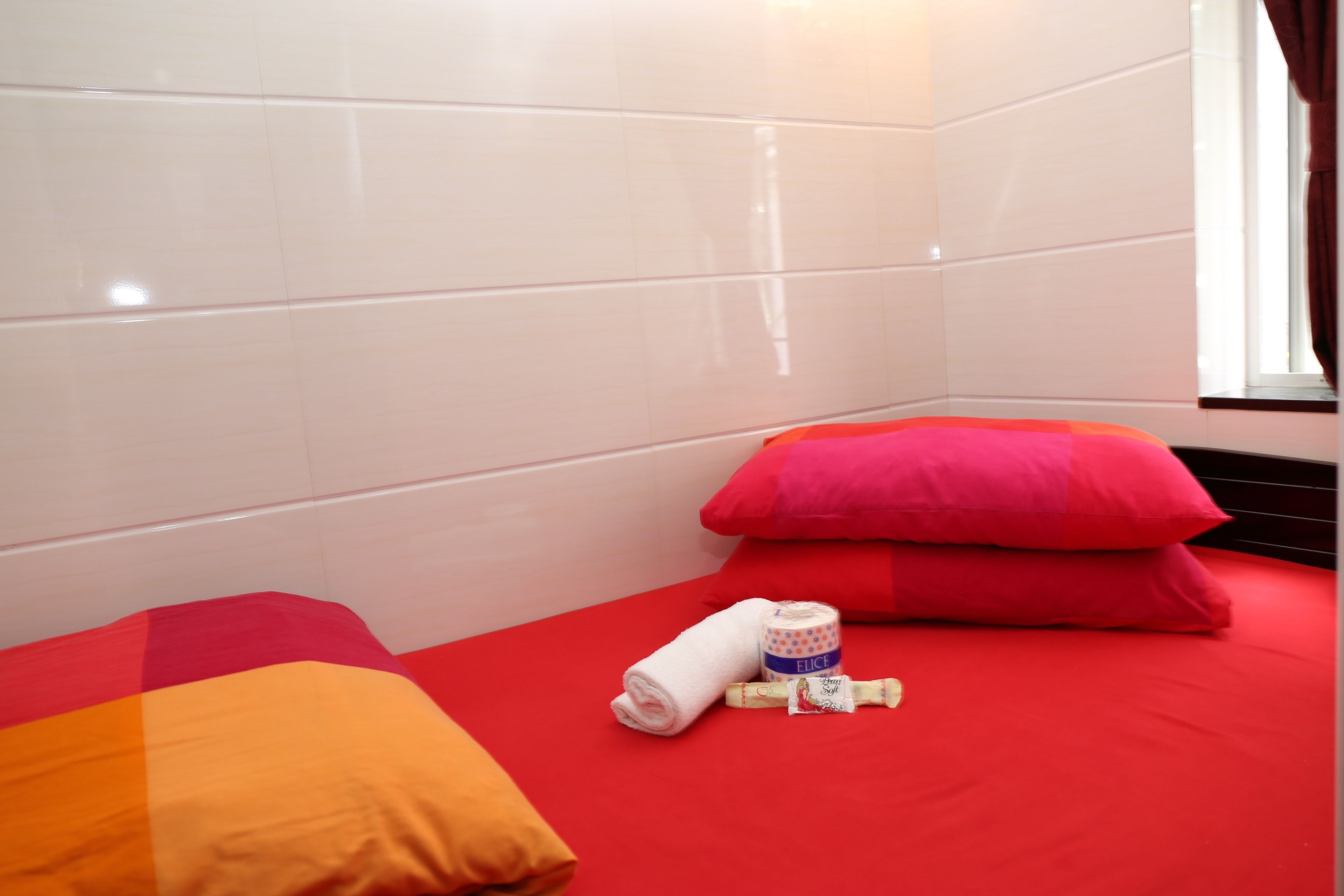 Une petite pièce, bien rangée, avec une couverture de lit rouge, deux oreillers, une serviette blanche enroulée et une bouteille de lotion.