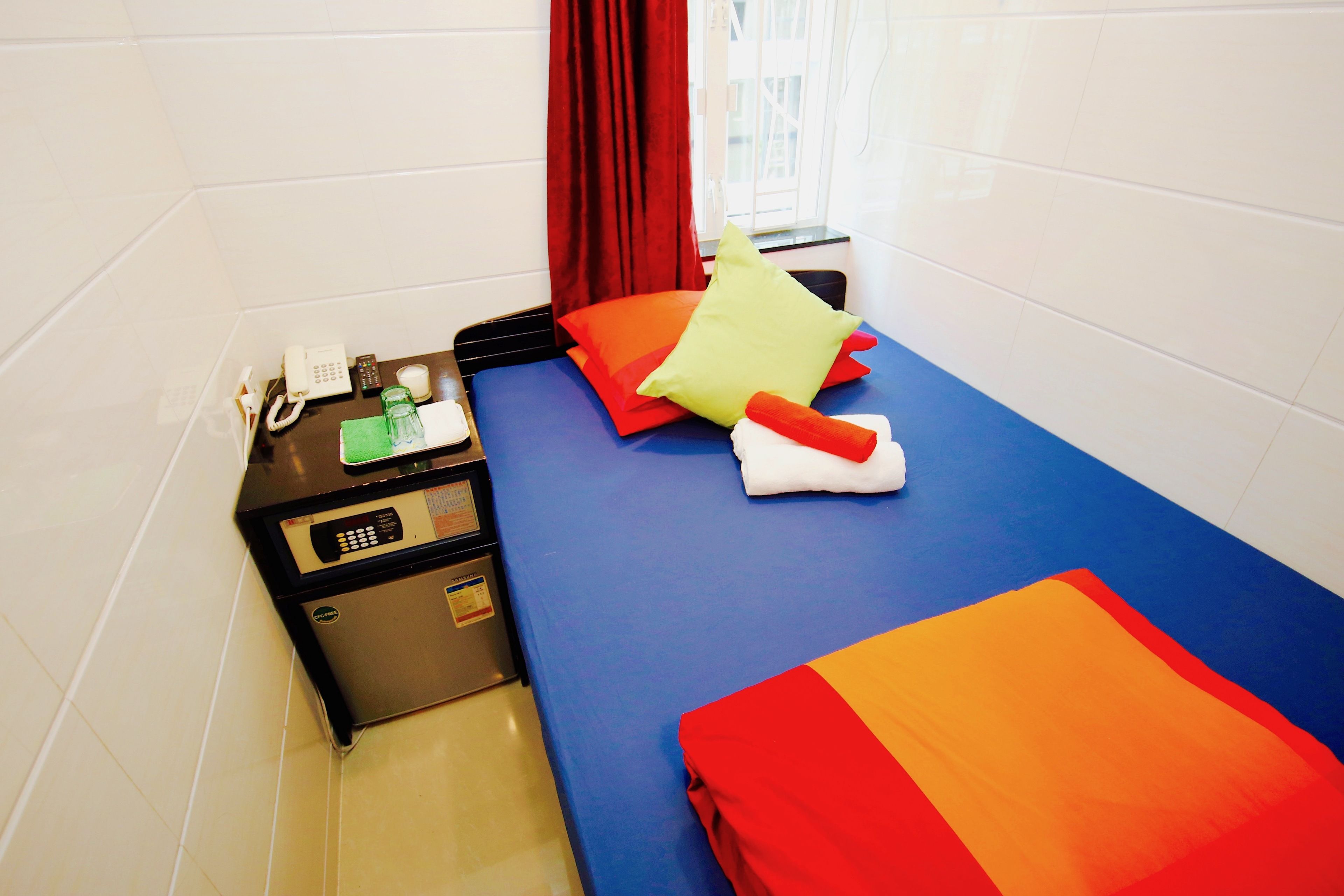 A hotel room with a bed, a desk with a phone, a small fridge, and a window with red curtains.