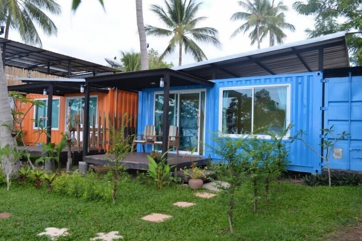 A blue shipping container house with a porch and a small garden area.