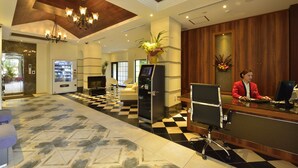 Zona de recepción de un hotel con mostrador de check-in, máquina expendedora, área de descanso con sofá y una planta decorativa.