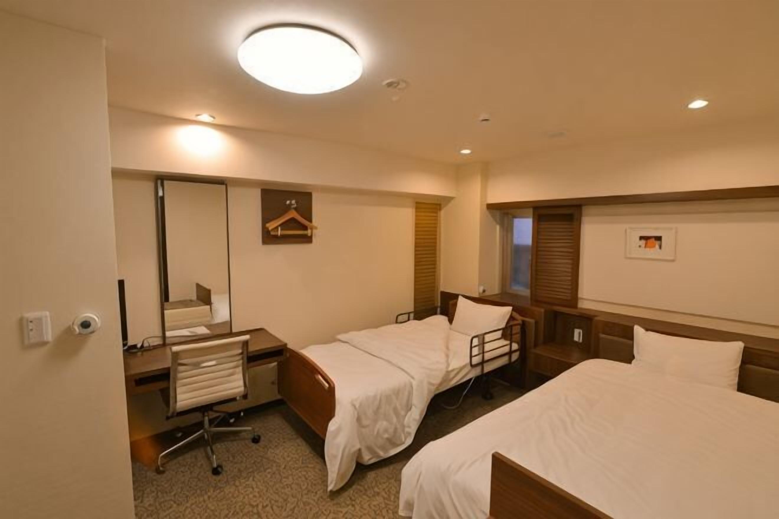A hotel room with two beds, a desk, a chair, a mirror, and a window.