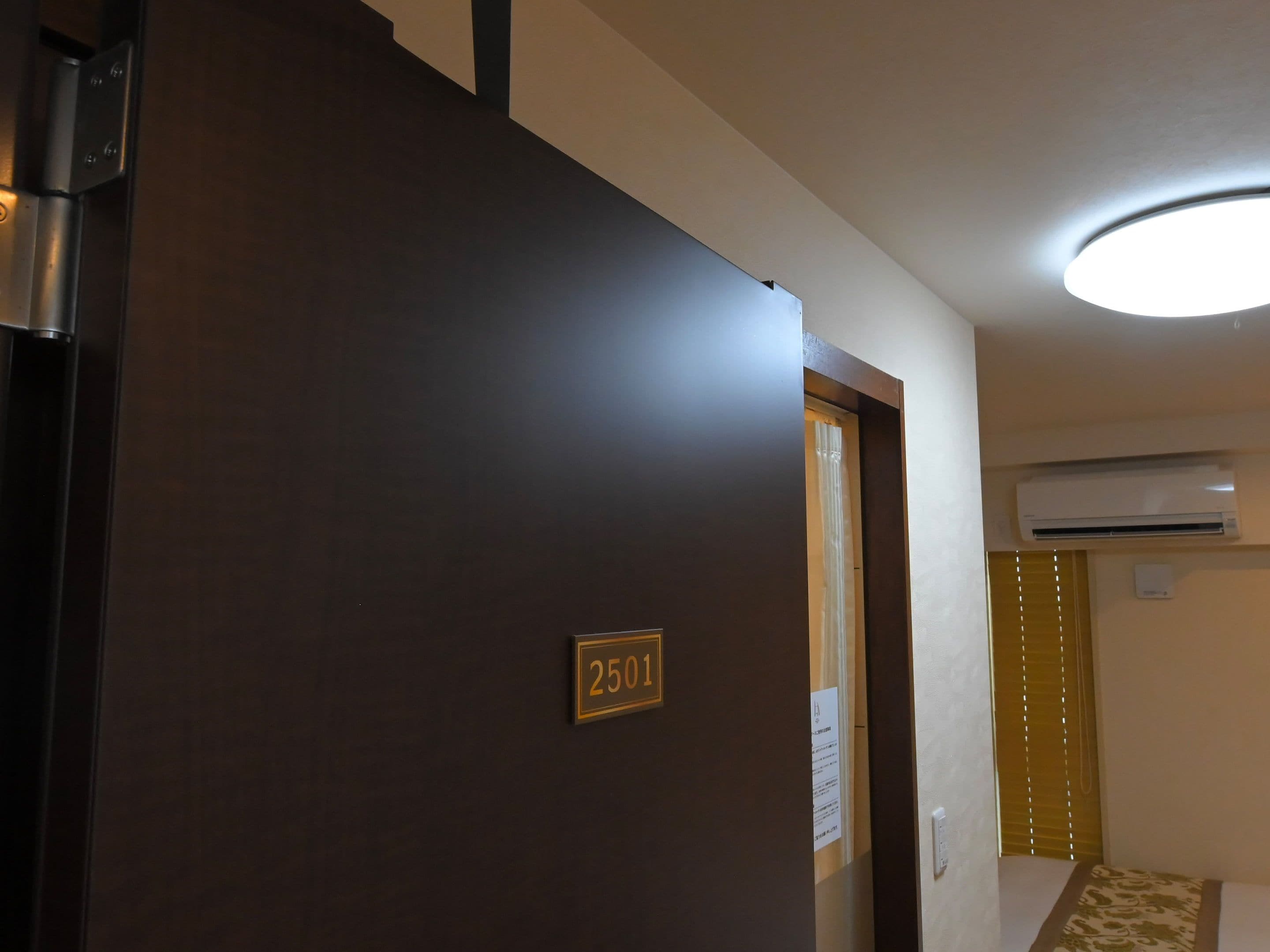 A hotel room entrance with a door labeled '2501,' an air conditioning unit mounted on the wall, and a ceiling light fixture.