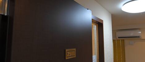 A hotel room entrance with a door labeled '2501,' an air conditioning unit mounted on the wall, and a ceiling light fixture.