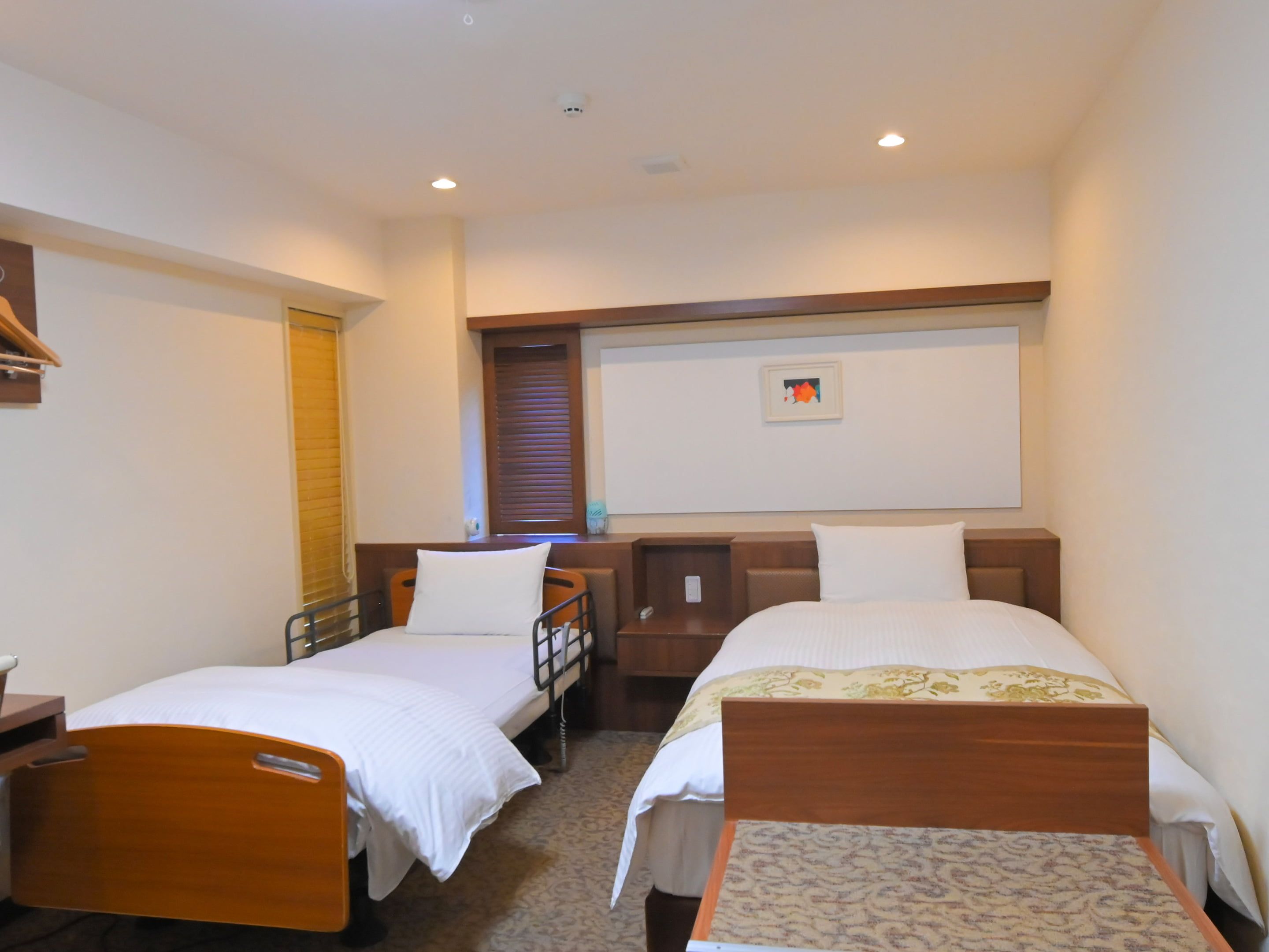 A hotel room with two single beds, a wooden desk, a small chair, a bedside table, and a window with blinds.