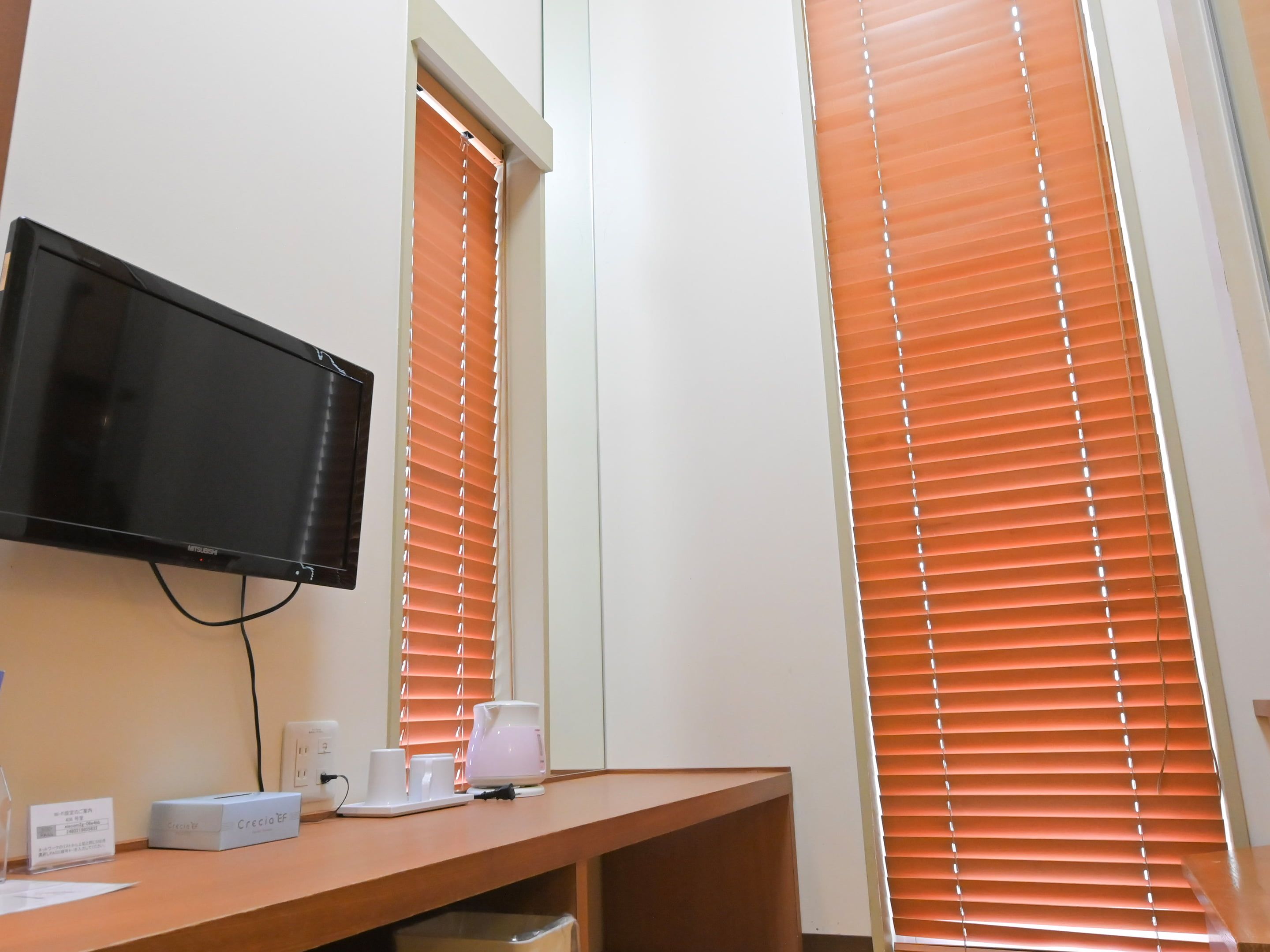 A hotel room with a flat-screen TV mounted on the wall, a wooden desk with a coffee maker, cups, and a box labeled 'Crecia EF'.