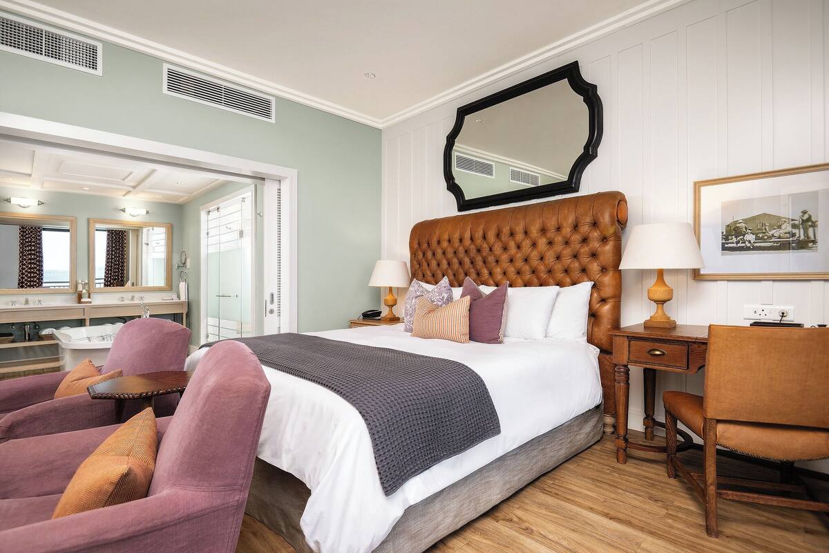 Ein Schlafzimmer mit einem großen Bett, einem Holz-Kopfteil, zwei Nachttischlampen, einem Schreibtisch mit Stuhl und einem Spiegel an der Wand.
