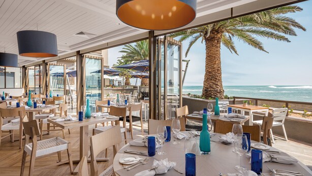 Un restaurant avec des tables dressées pour le service, de grandes fenêtres et une vue sur l’océan.