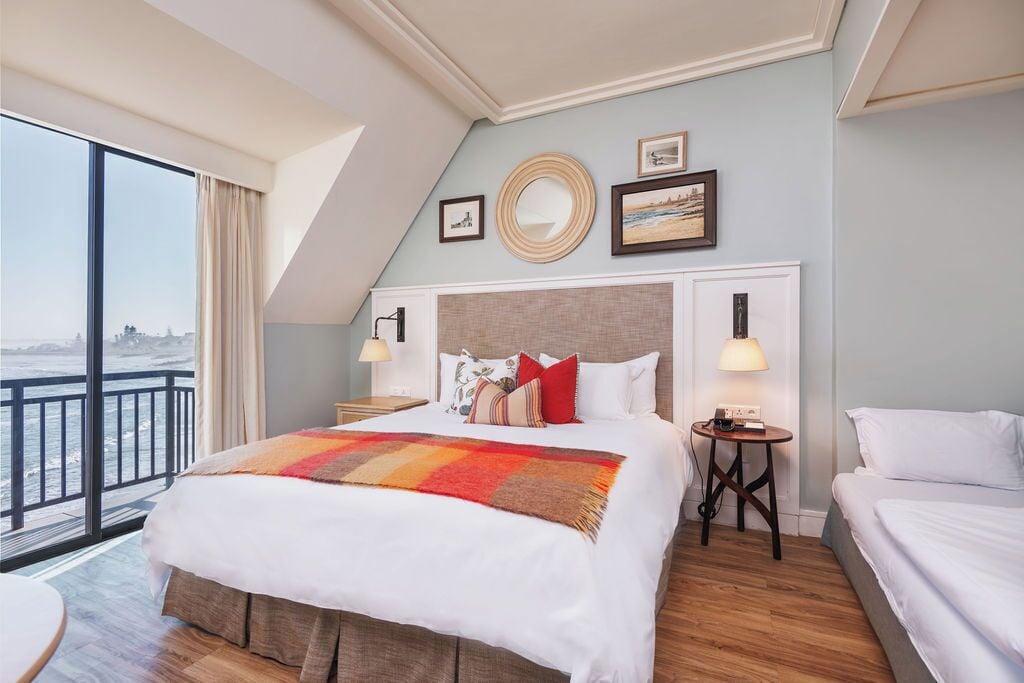 A hotel room with a large bed, a smaller bed, a round mirror, framed pictures, and a view of the sea.