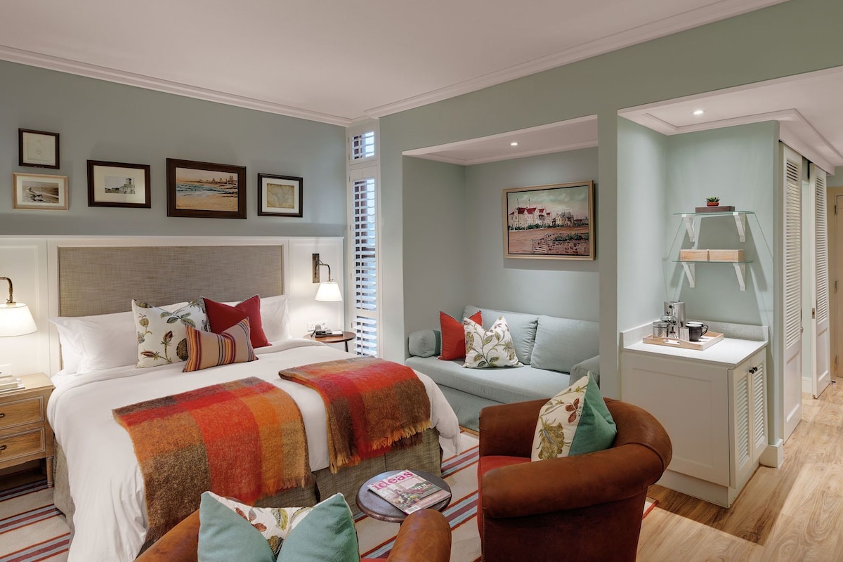 Habitación de hotel con cama, sofá, sillón, escritorio con lámpara y un cuadro en la pared.
