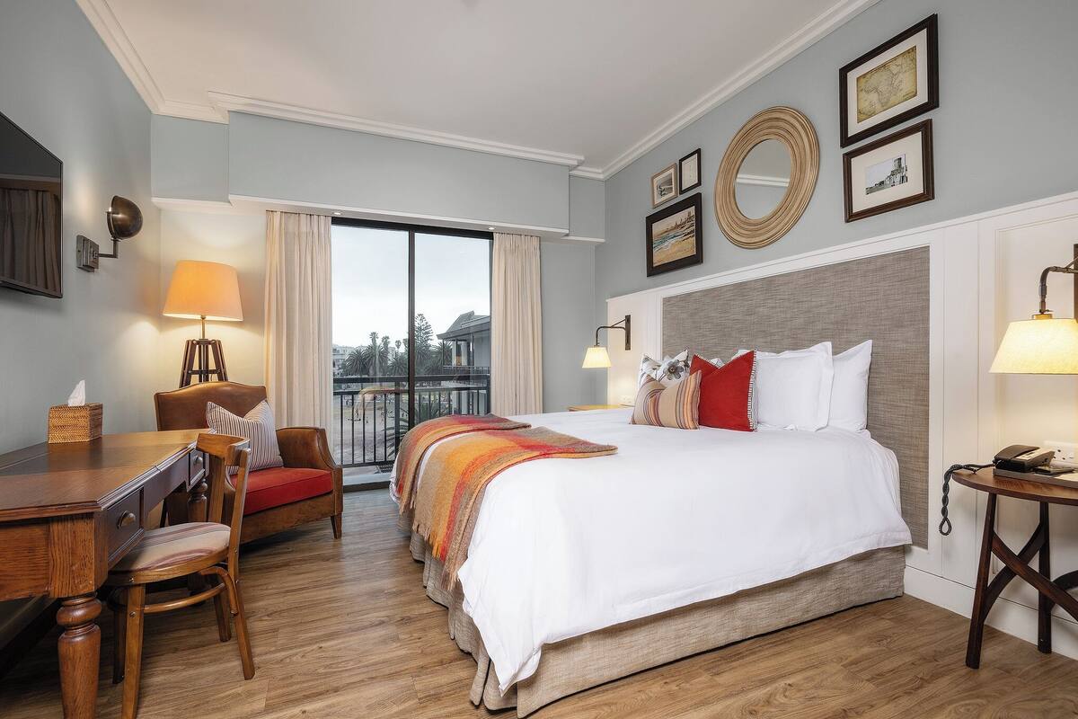 Ein Hotelzimmer mit einem großen Bett, einem Esstisch, einem Stuhl, einem Balkon mit Vorhängen und gerahmten Bildern an der Wand.