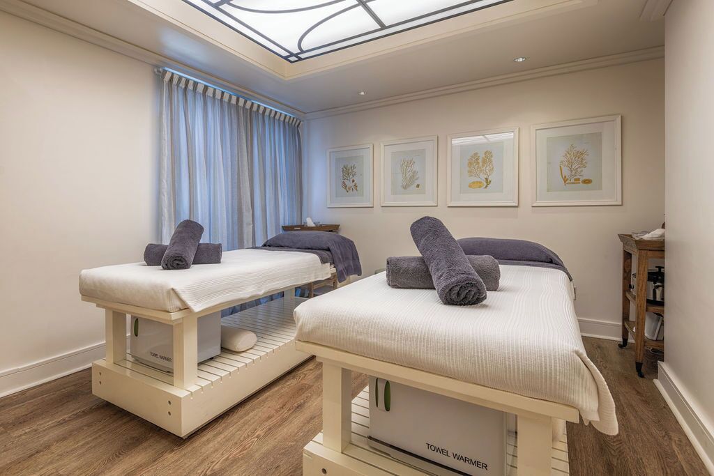 A room with two beds, a skylight, a window with curtains, and framed botanical prints on the wall.