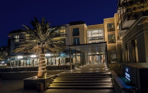 Entrada de hotel com palmeira, área de estar ao ar livre e um caminho bem iluminado que leva ao edifício.