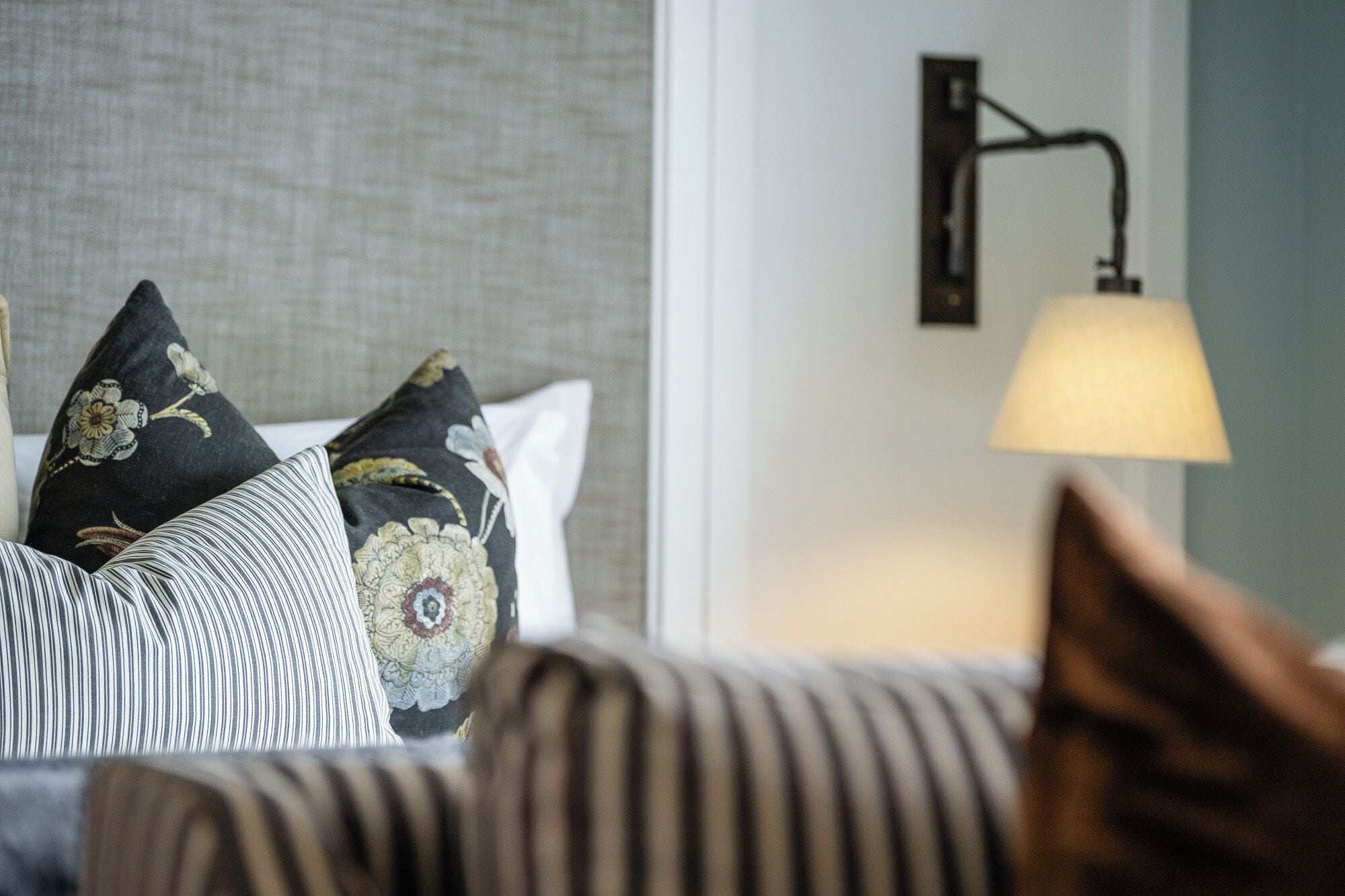 A hotel room with a bed featuring striped bedding, a headboard, and decorative pillows. There is a wall-mounted lamp on the right side.