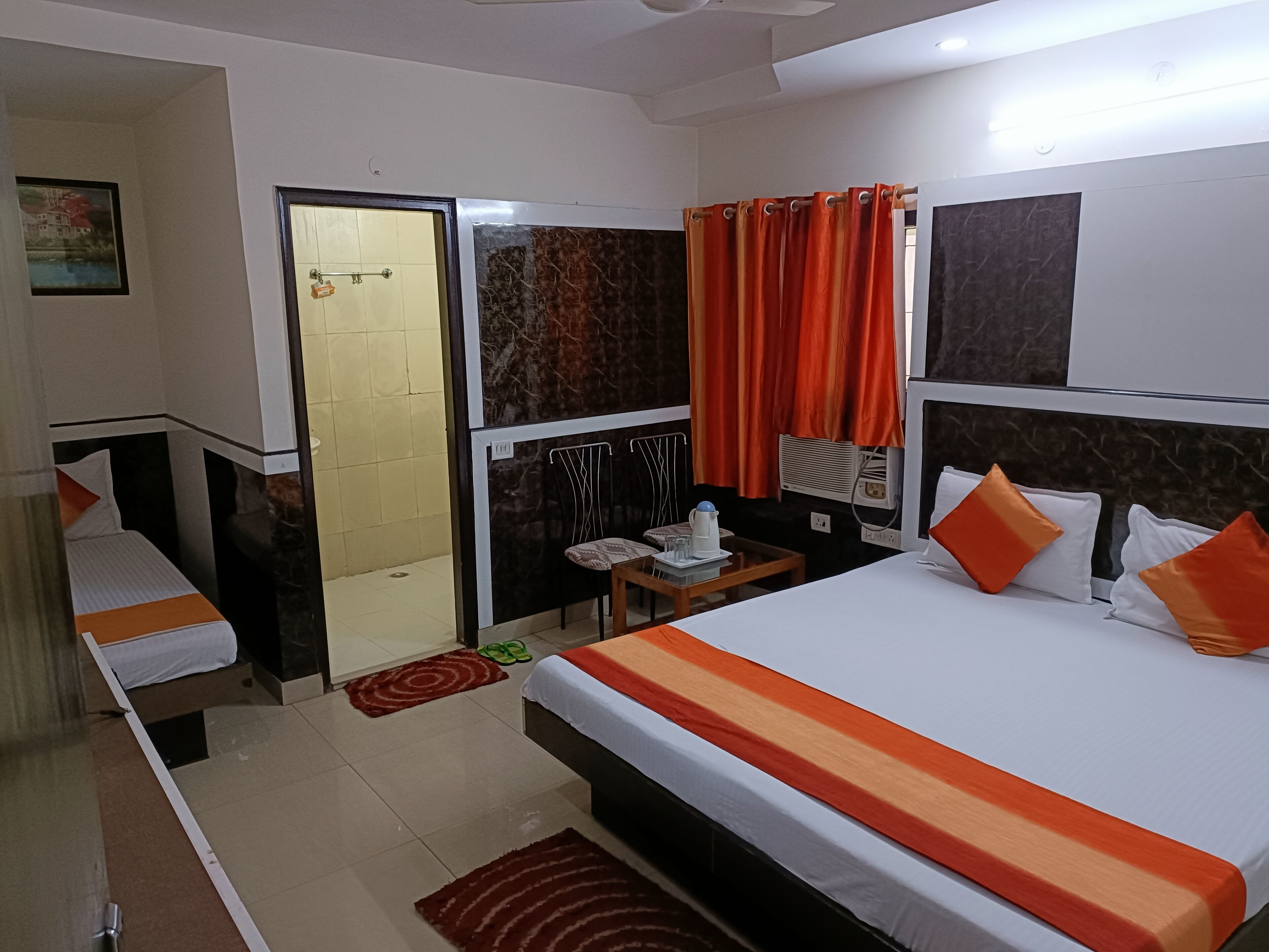 A hotel room with two beds, a small table, a chair, a wardrobe, and a bathroom with a shower and toilet.