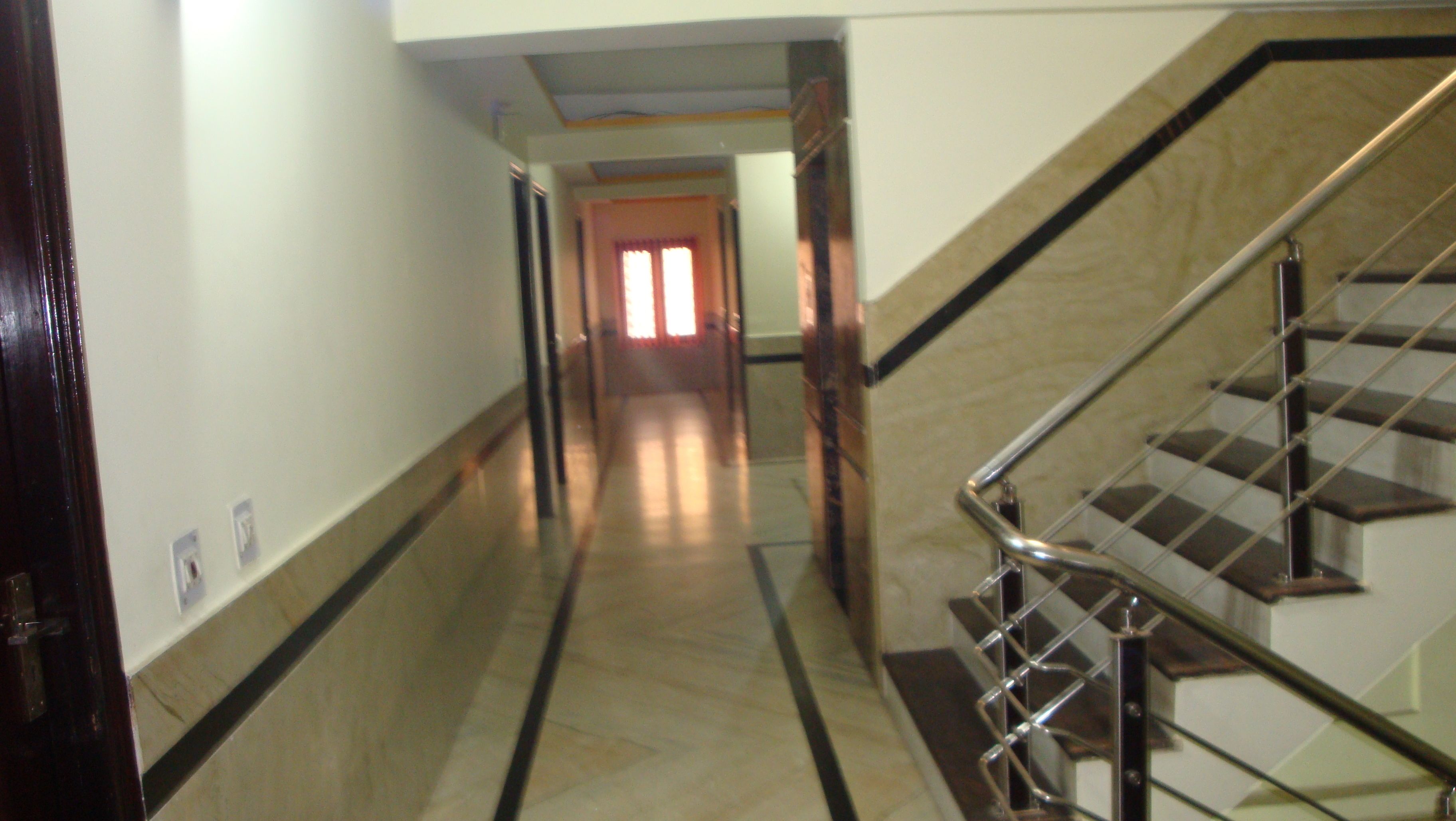 Un couloir avec un escalier sur la droite, menant à l’étage supérieur. Le sol est recouvert de carreaux clairs et foncés disposés en motif.
