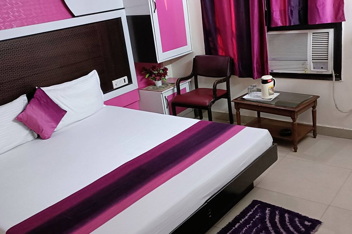 A hotel room with a bed, a desk, a chair, a window with curtains, and an air conditioning unit.