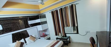 A hotel room with a bed, a desk, a chair, a window with curtains, a ceiling fan, and a marble-patterned wall.