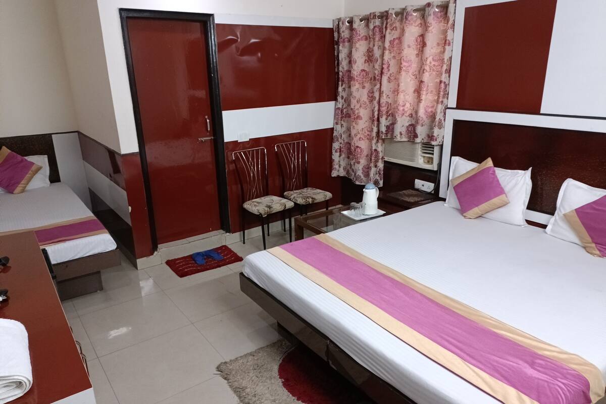 A hotel room with a large bed, two smaller beds, a desk, a chair, a red door, and a window with curtains.