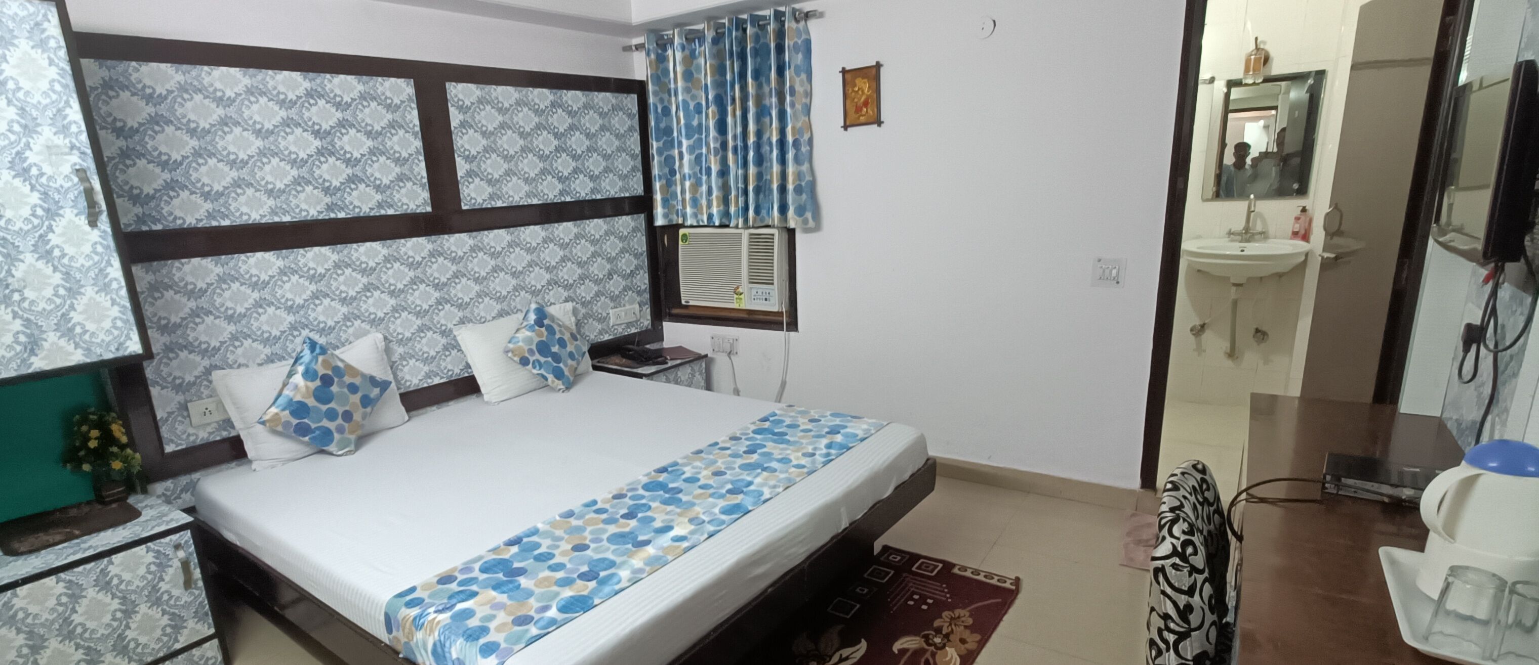 A bedroom with a bed, a bedside table, a nightstand, a window with a curtain, a wall-mounted air conditioner, a mirror, a sink, and a cabinet.
