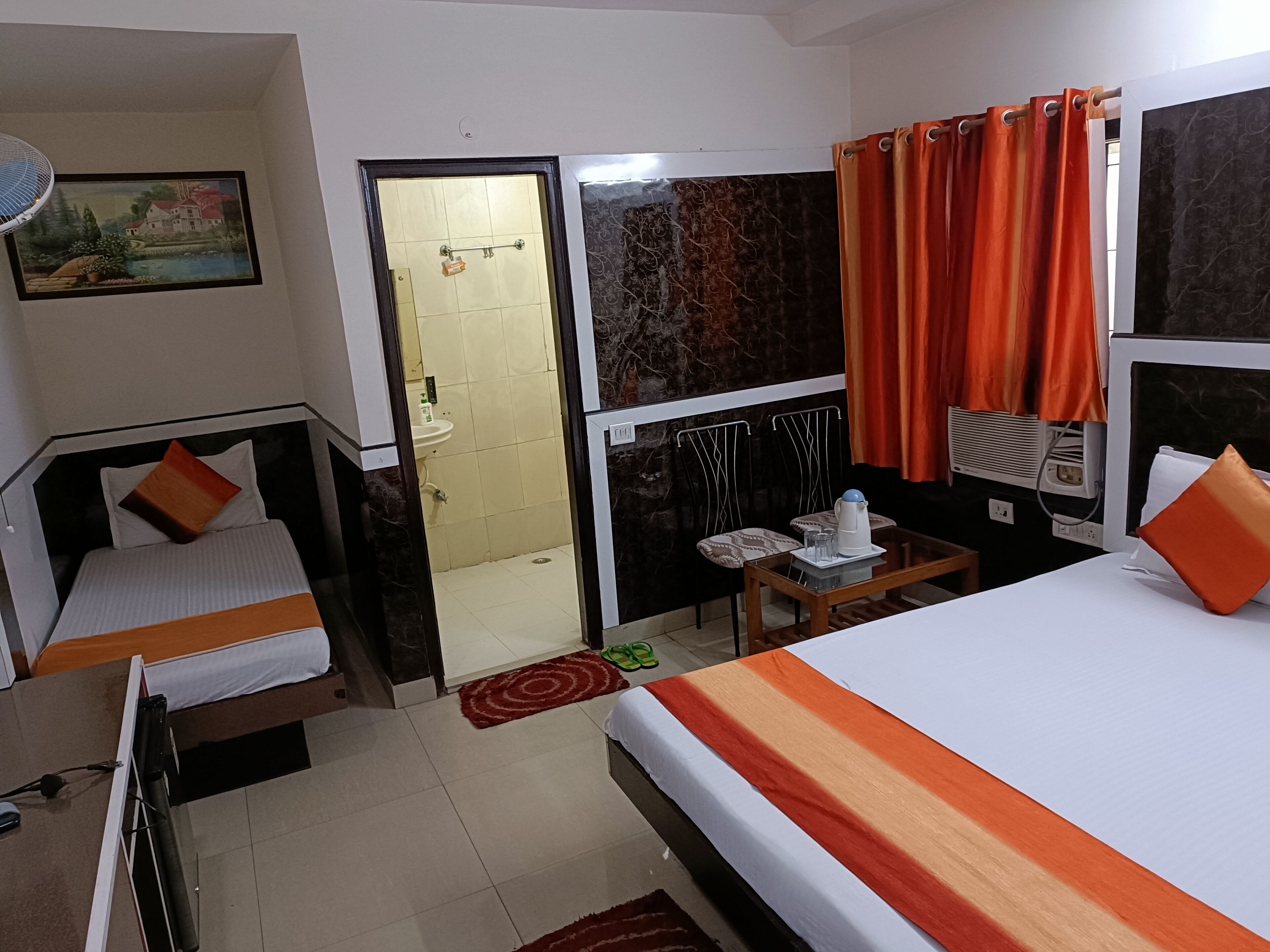 Une chambre d’hôtel avec deux lits, une petite table avec une bouilloire, une chaise, une fenêtre avec des rideaux et un tableau au mur.