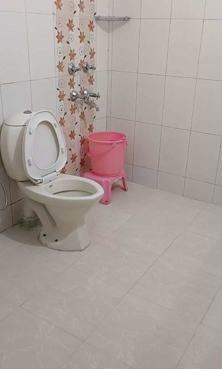 A bathroom with a toilet, a pink bucket, and a shower with a floral pattern on the wall.
