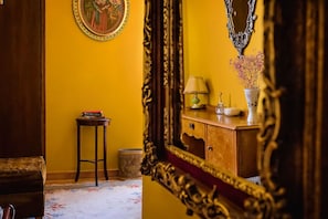 A room with a yellow wall, a wooden cabinet, a small table with books, a lamp, and a vase with dried flowers. - Athens Quinta - Hostel (Athens)