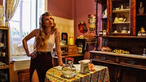 A woman standing in a kitchen with a table set for tea, surrounded by various kitchen utensils and decorative items.