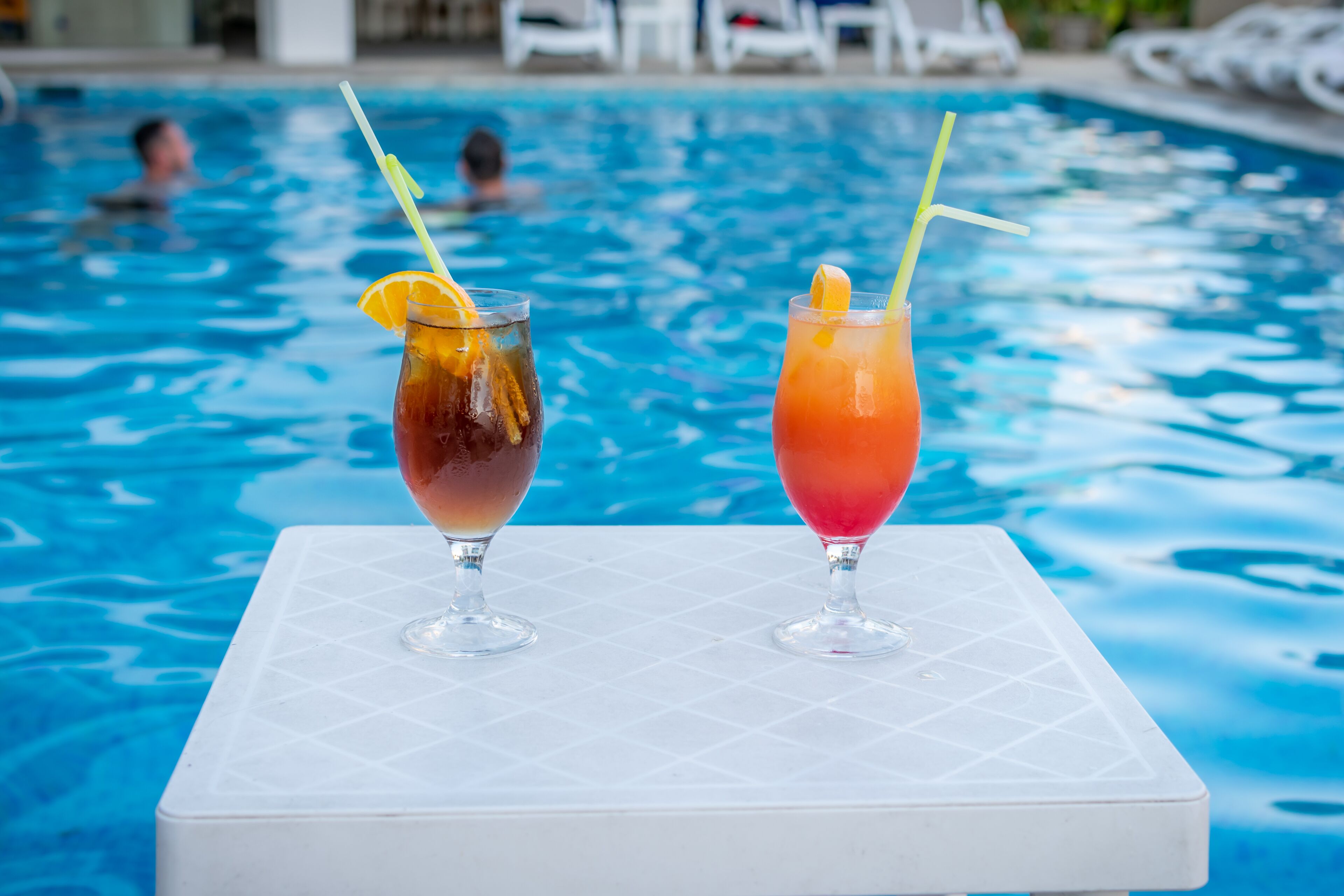 Two drinks on a white table by a pool with people in the background.