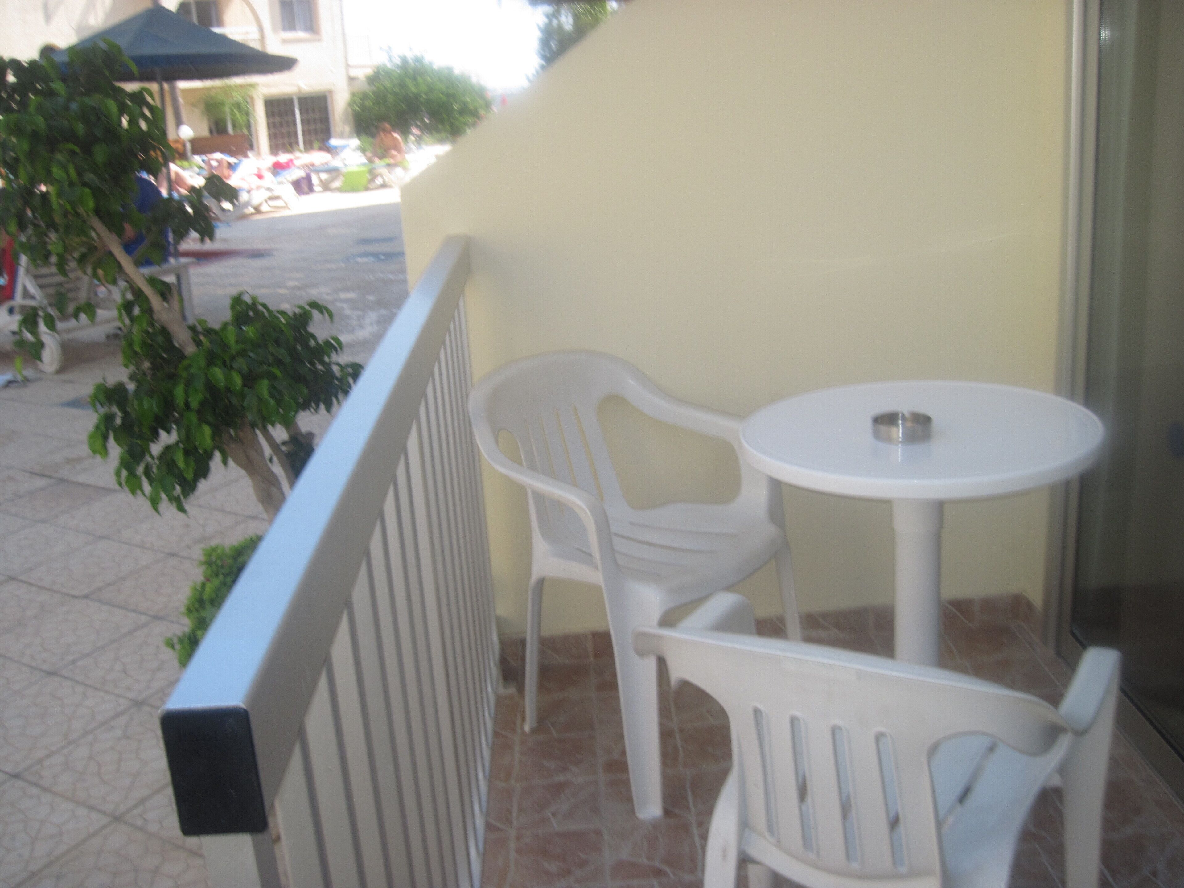 Un balcon avec une chaise et une table en plastique blanc, un sol carrelé et un immeuble en arrière-plan.