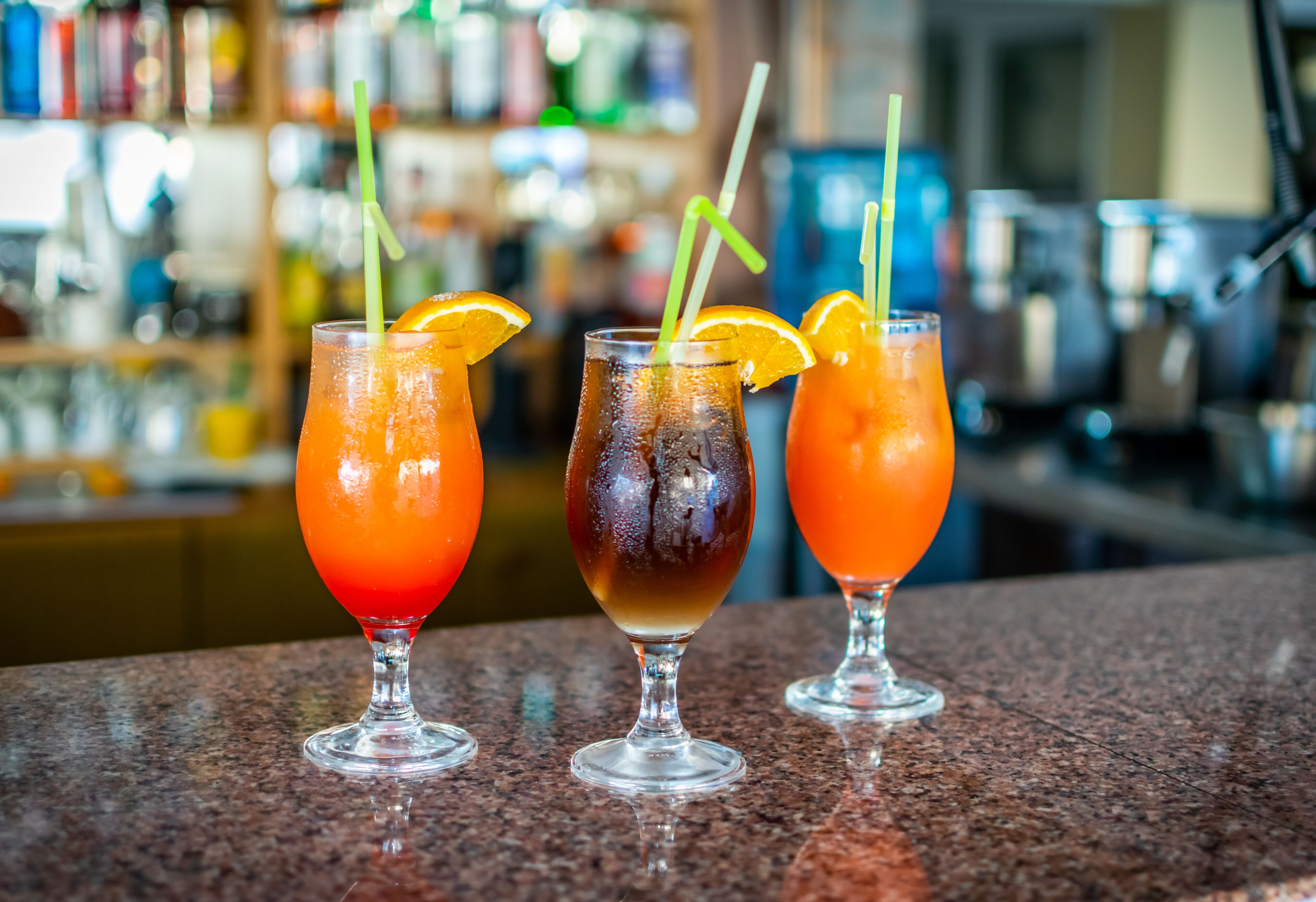 Tres vasos de bebidas sobre una barra, con un fondo desenfocado de estantes y botellas.