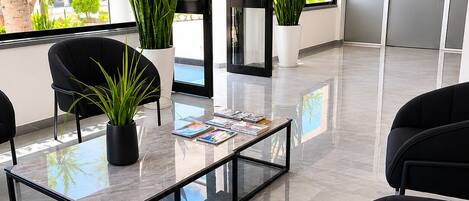 A modern waiting area with a glass coffee table, black chairs, potted plants, and a view of the outdoors.