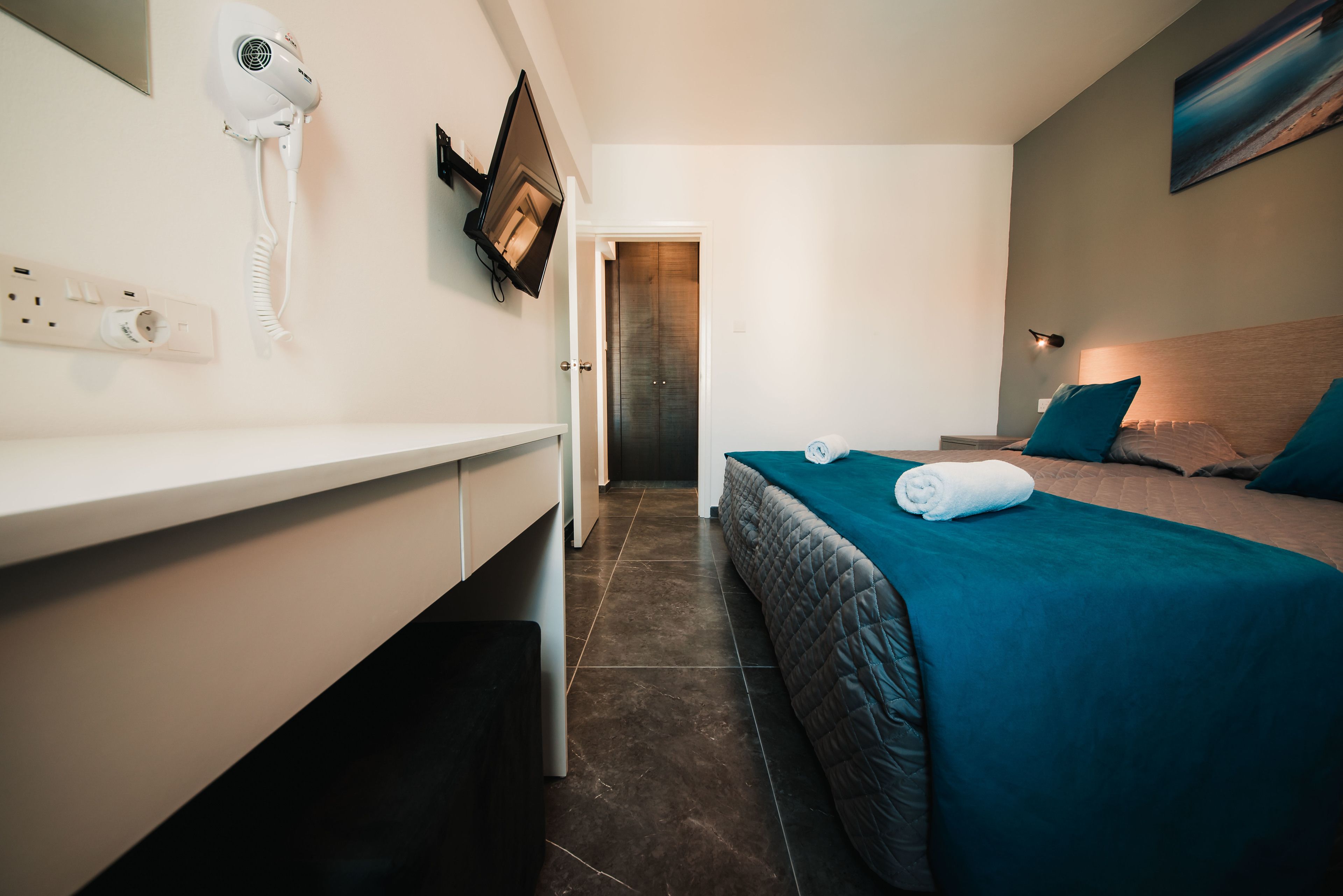 Habitación de hotel con cama, televisor montado en la pared, secador de pelo y baño con lavamanos y espejo.