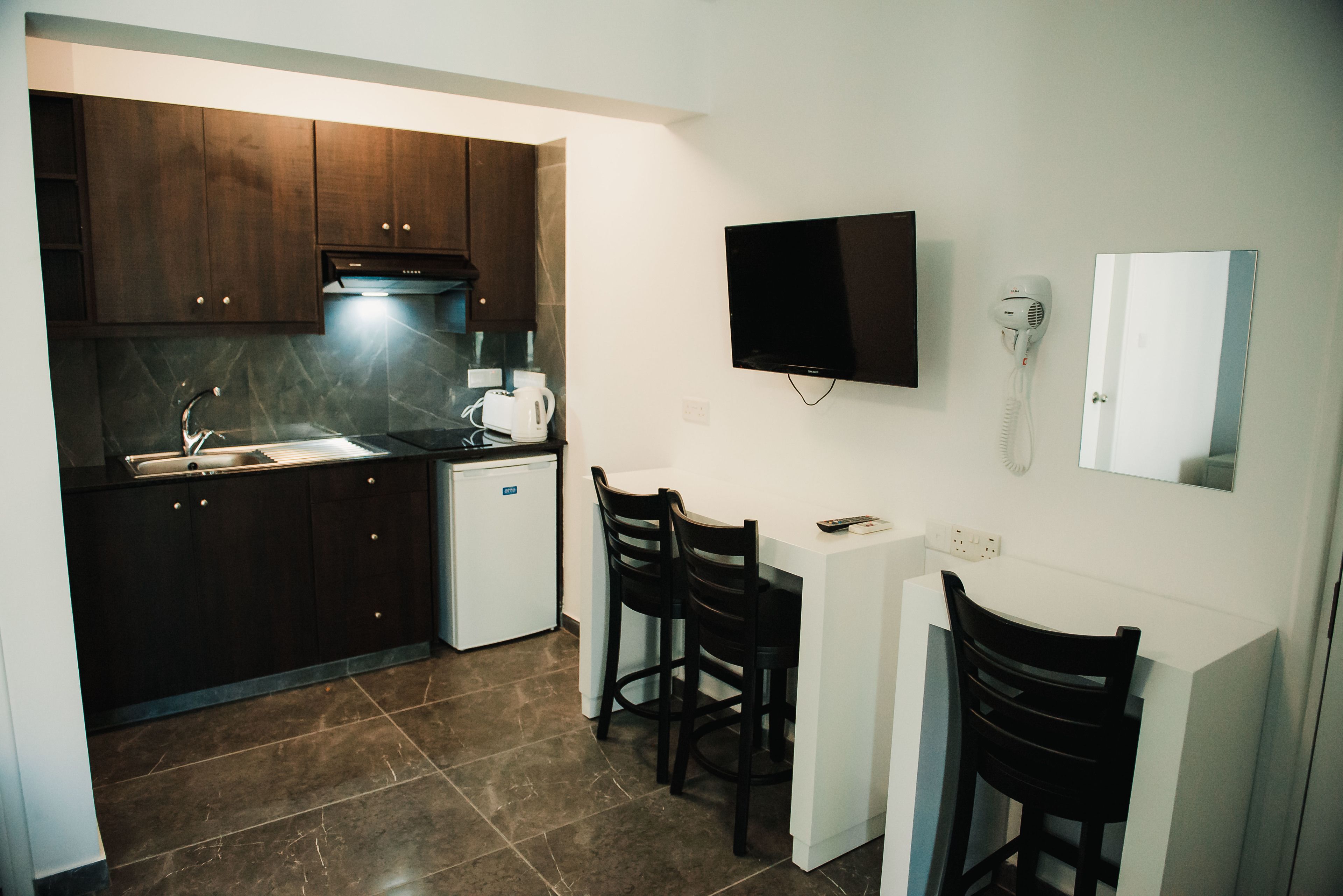 Una cocina compacta con armarios de madera oscura, un horno empotrado y un refrigerador pequeño. Hay una encimera blanca con una cafetera y un secador de pelo en la pared. Dos sillas negras están colocadas frente a una mesa blanca pequeña.