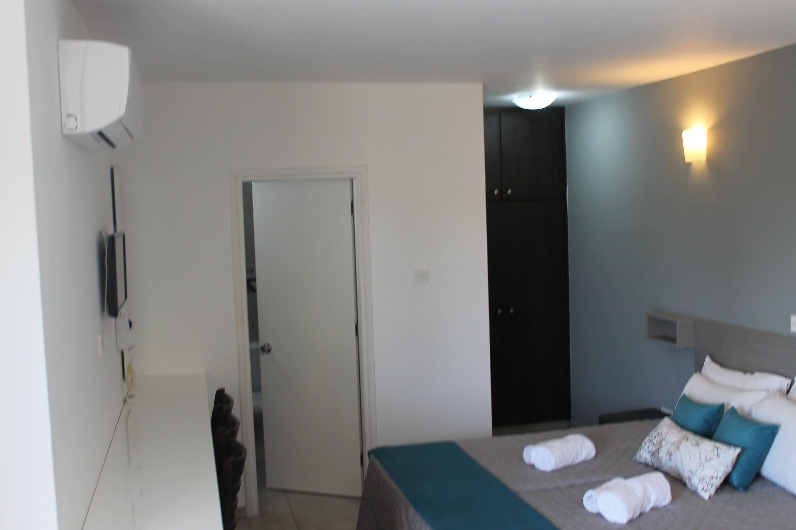 A hotel room with a bed, a door, a wall-mounted light, and a small shelf.