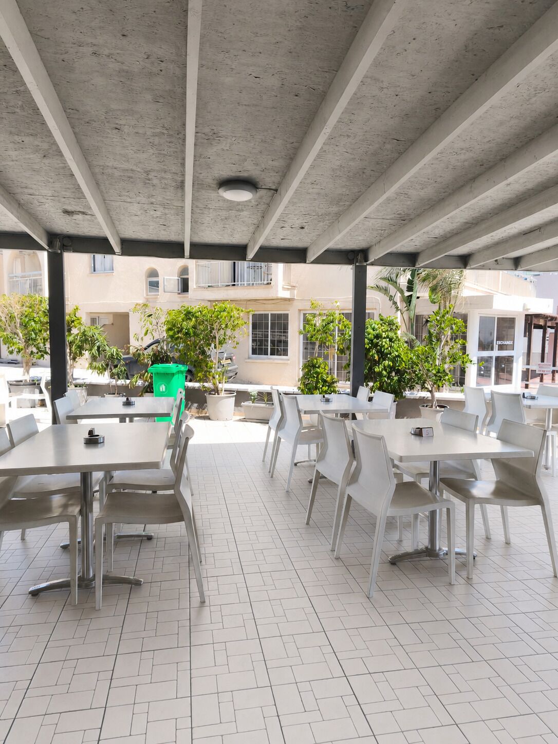 An outdoor patio area with white tables and chairs, a concrete ceiling, and potted plants.