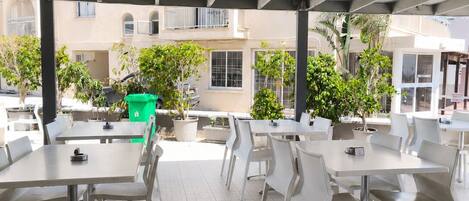 An outdoor patio area with white tables and chairs, a concrete ceiling, and potted plants.