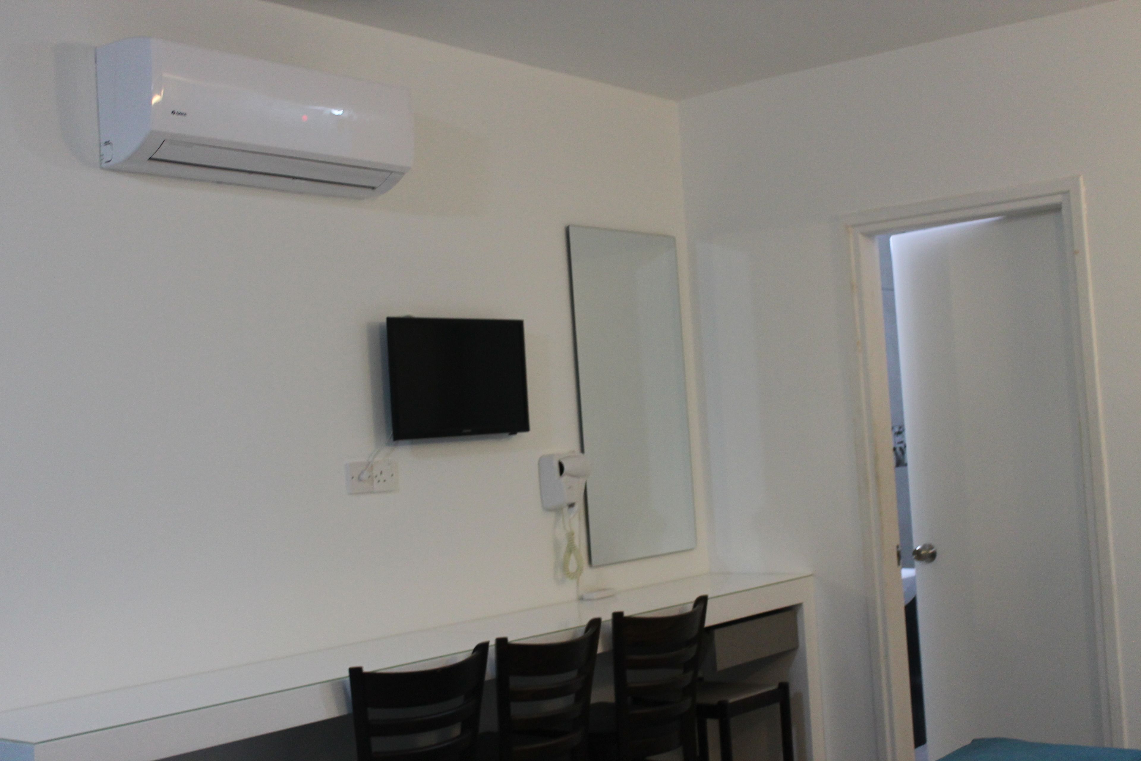Une pièce avec un climatiseur blanc, un téléviseur à écran plat fixé au mur, un grand miroir et une porte donnant sur une autre pièce.