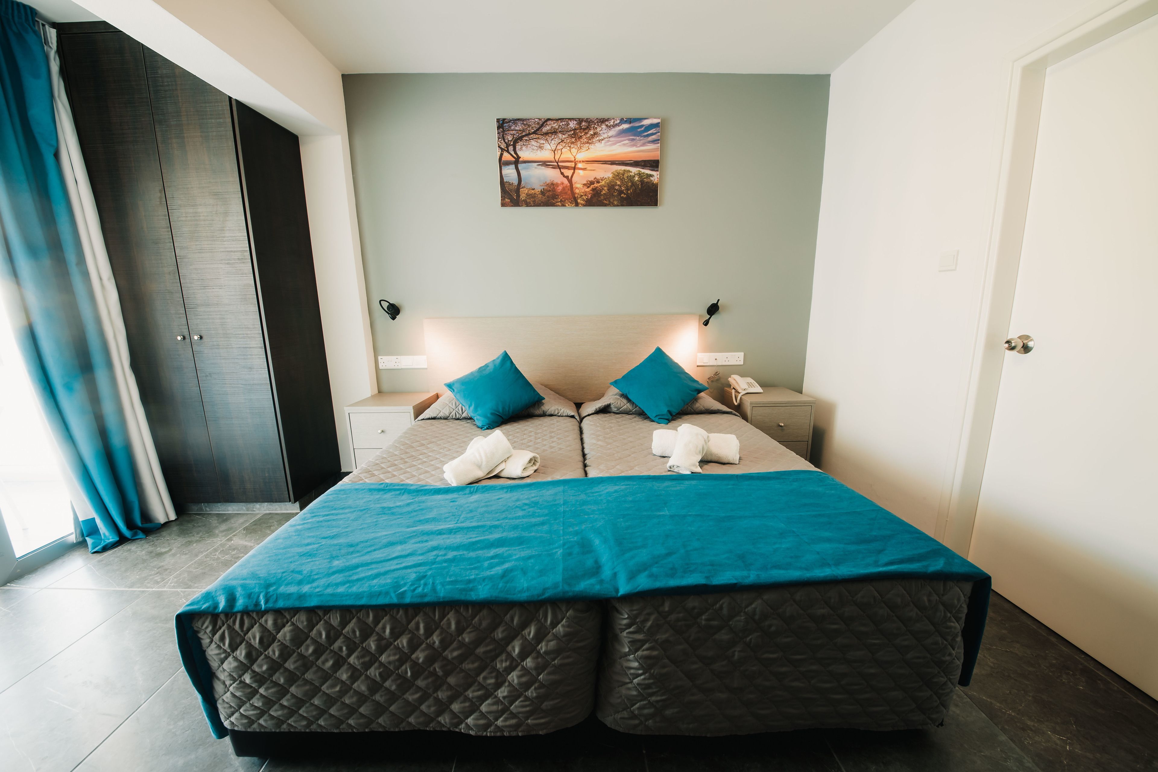 Cama doble con cabecera acolchada, dos almohadas y una manta color turquesa. Se ve una mesita de noche con una lámpara, un cuadro en la pared y una puerta a la izquierda.
